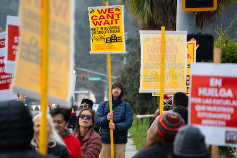 people on strike with picket signs. at focus in the center is a man with a sign that says we can't wait. invest in the schools our students deserve. fully staffed schools. stability for our students. living wages for educators