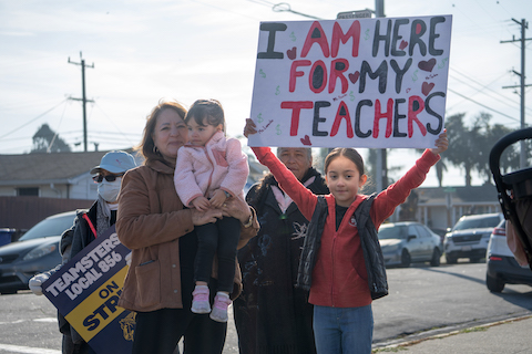 a girl holds a sign that reads i am here for my teachers. with her are a woman holding a younger girl and an older woman. near them is a person with a ballcap, sunglasses, surgical mask and sign that reads teamsters local 856 on strike
