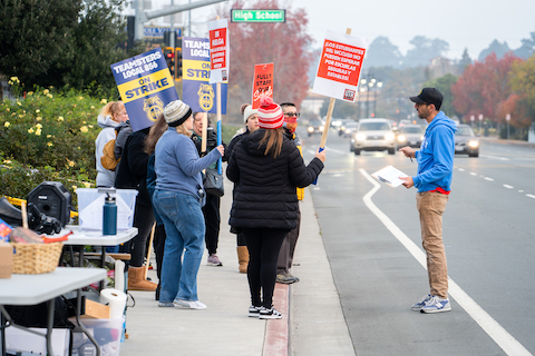 people on strike with picket signs standing on a sidewalk. two signs say teamsters local 856 on strike. one sign says fully staff our schools. two signs are in Spanish. a street sign says High School. a man with a clipboard is standing in the street