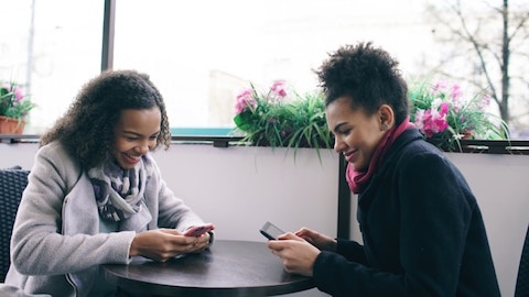 two smiling young women sitting across from each other at a table both looking at their phones