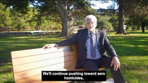 latino man with white hair and goatee wearing a suit and tie. he is sitting on a park bench surrounded by green grass with trees in the background. closed captioning shows him saying we'll continue pushing toward zero homicides