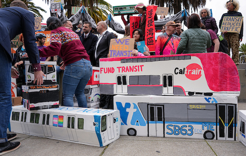cardboard decorations made to look like a bart train, a cal train car that says fund transit, and a VTA bus that says SB 63 for me. a crowd of people is in the background with one person holding a sign that says we need transit and another with a sign that says transit reduces traffic