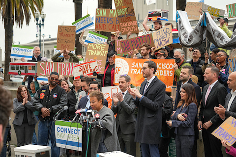 black woman at lectern with news station microphones and sign that says connect bay area. several people are standing behind her many with signs that say things like we need transit, san francisco transit riders, save my bus, say no to bart cuts that could close san bruno station, strong transit strong bay, fund transit now