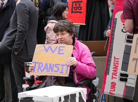woman in electric wheelchair holding sign that says we need transit. a crowd is standing around her including one person with a sign made to look a cal train car with fund transit written on it