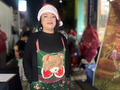 latina woman wearing a santa hat and a green christmas themed apron with a large teddy bear head wearing a red bow tie on it