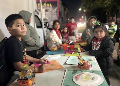 latino children standing at a table outside at night with colored paper, markers and other art supplies. santa is drawn on a paper plate