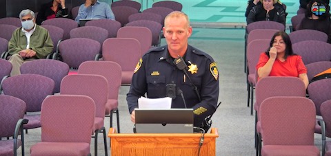 white man in police uniform at lectern with a few people seated in audience behind him