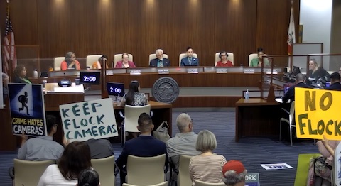 a scene from a richmond city council meeting shows signs in the audience that read crime hates cameras, keep flock cameras, and no flock