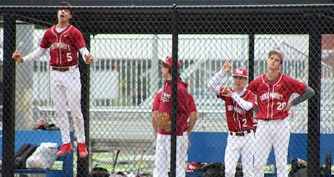 four high school baseball players behind a chain link fence. one has jumped in the air. his feet are far off the ground and his hands are on the fence. another has his hands on his hips and looks displeased